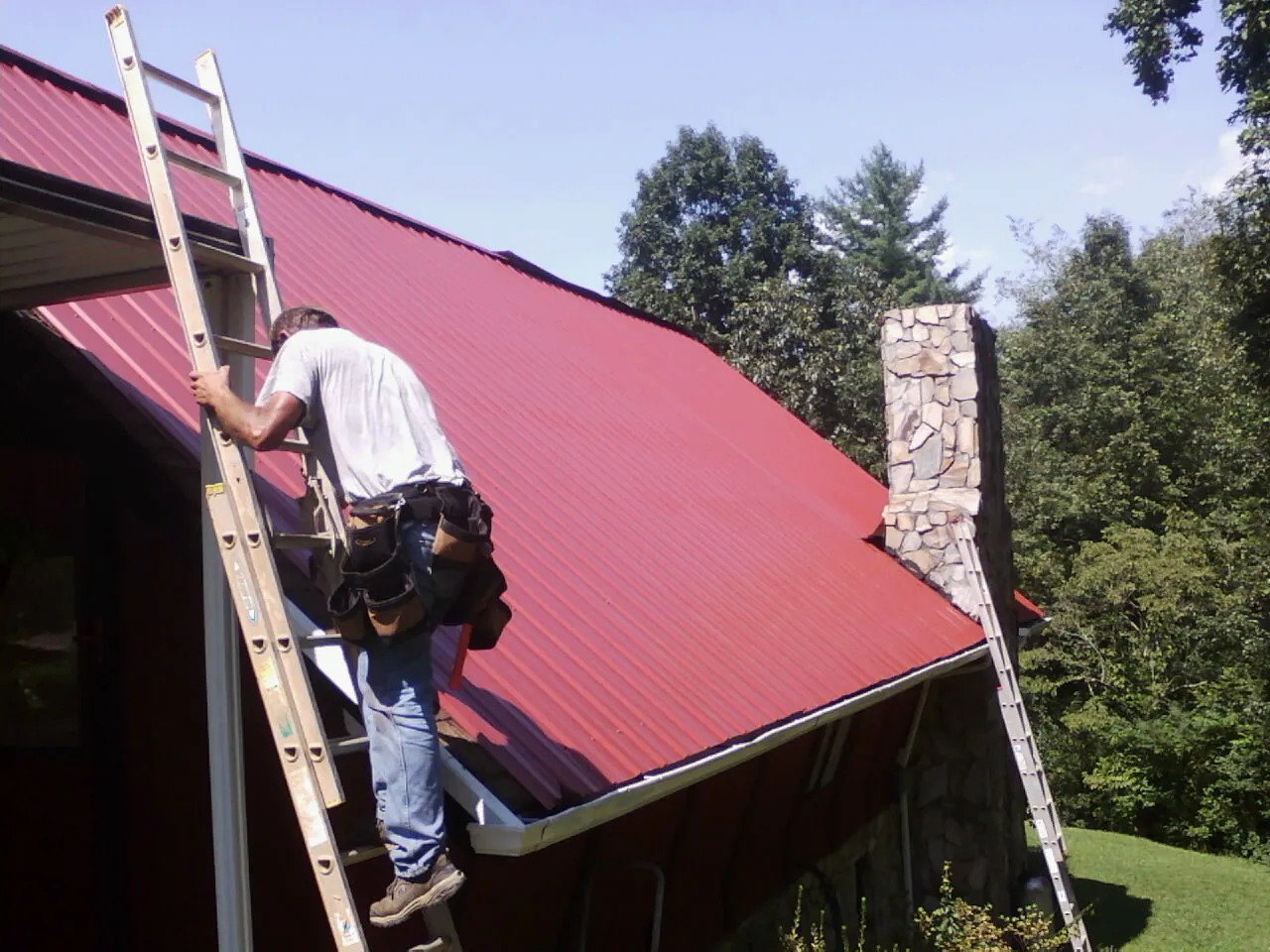 Roofer on a ladder beside a red metal roof and stone chimney — Soffit & Fascia Repair by Roofing Brothers in Clarksboro